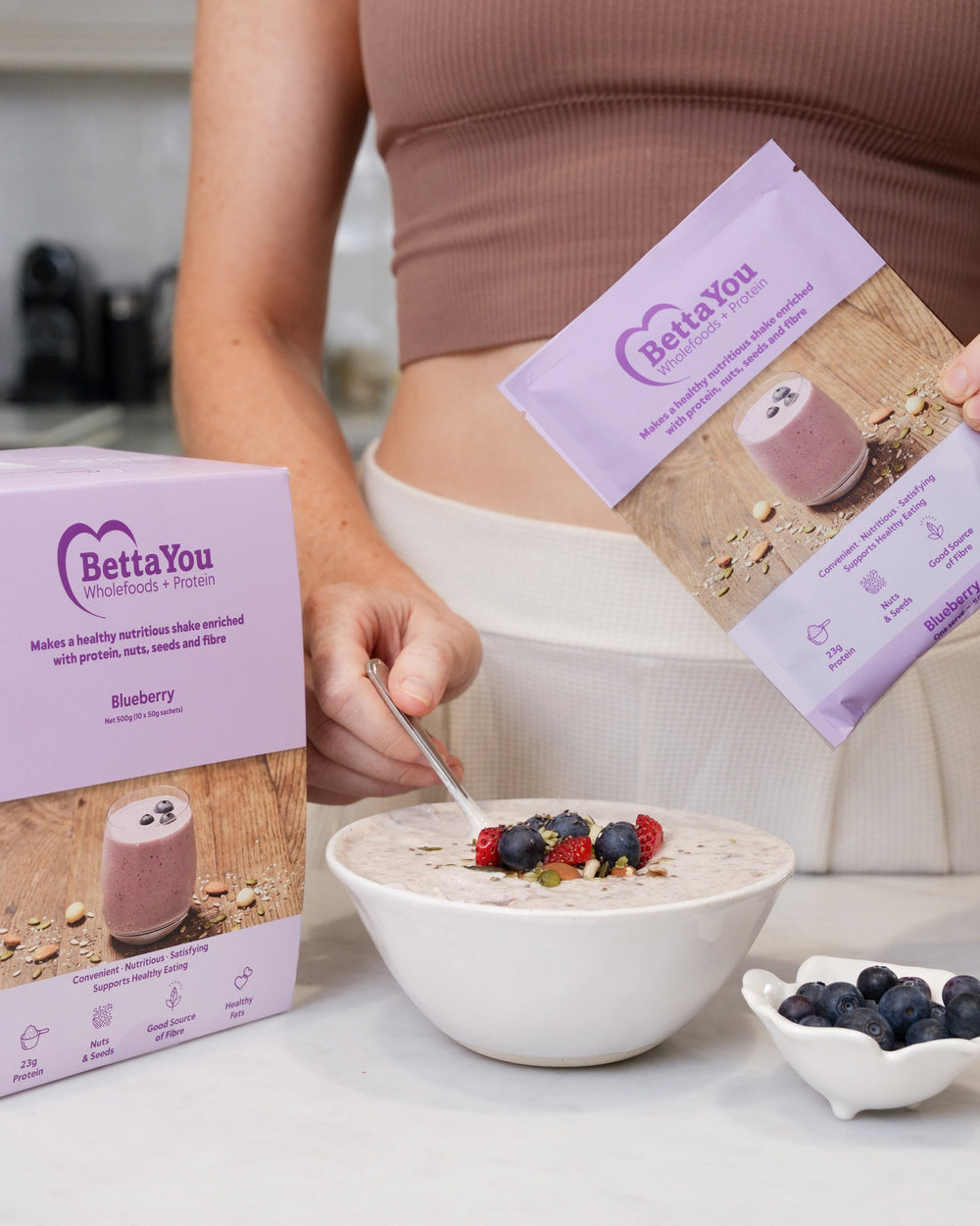 Person eating a bowl of yoghurt with bettaYou protein blueberries, surrounded by Betta You products.